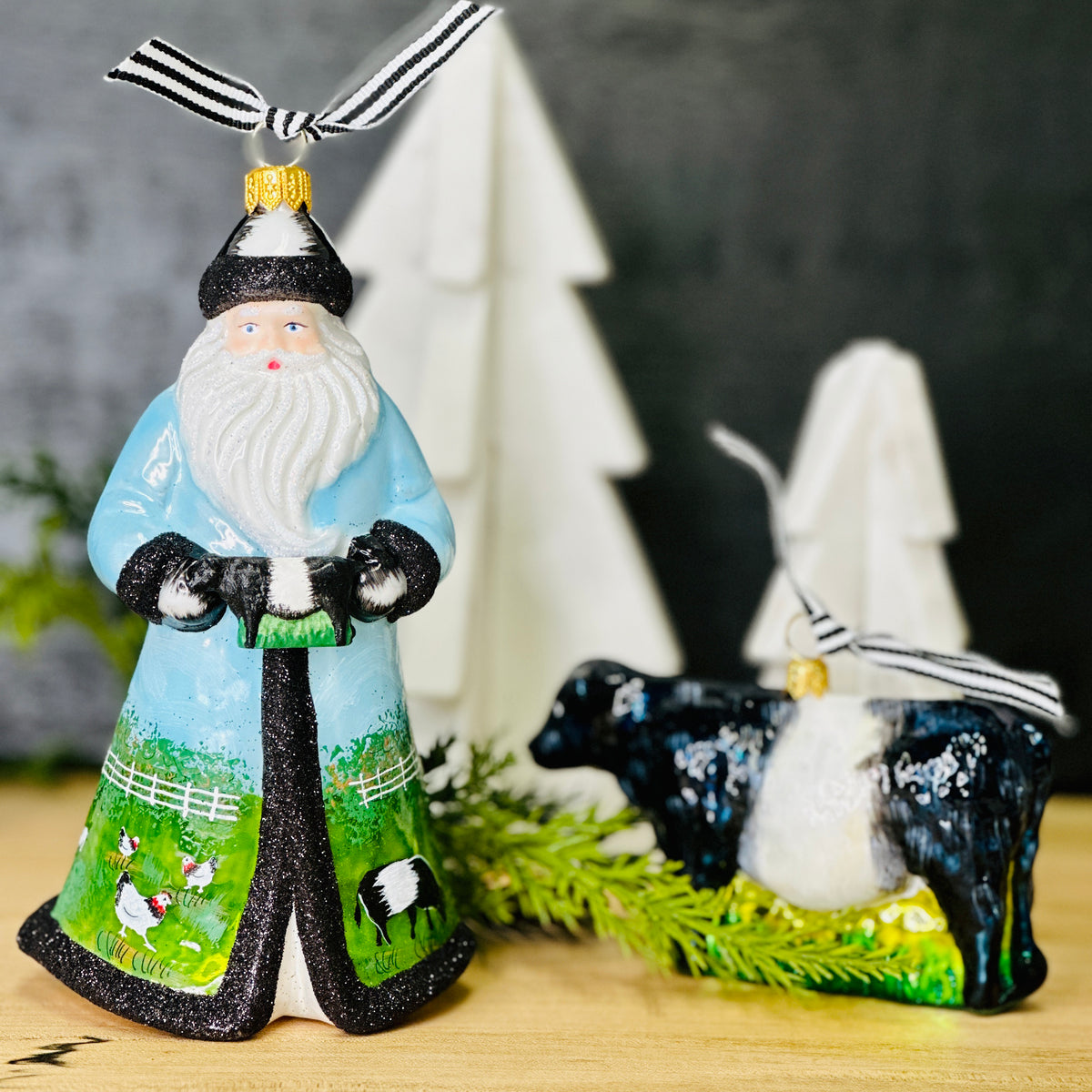 A luminous, hand-painted FEARRINGTON SIGNATURE COLLECTION - HANDMADE BELTIE ORNAMENT from JOY TO THE WORLD COLLECTIBLES dangles by a black and white striped ribbon. Shaped like a cow with a glossy surface that catches the light, it features a black head and rear with a white midsection. This delightful piece exudes the spirit of Fearrington Village, perched on a green base resembling grass.