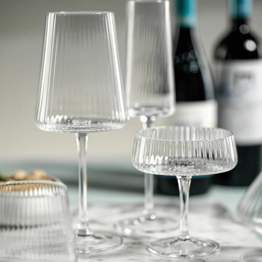 The FLUTED TEXTURED MARTINI GLASS by ZODAX stands empty against a white background, showcasing its wide, shallow bowl perched on a thin stem. Vertical ridges adorn the clear surface of the glass, adding texture to its elegant design. A flat, round base ensures stability.