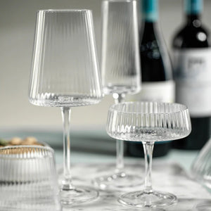The FLUTED TEXTURED MARTINI GLASS by ZODAX stands empty against a white background, showcasing its wide, shallow bowl perched on a thin stem. Vertical ridges adorn the clear surface of the glass, adding texture to its elegant design. A flat, round base ensures stability.