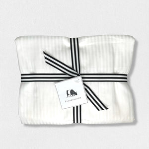 A neatly folded white fabric bundle, tied with a black and white striped ribbon, features a white tag illustrated with a small animal and the text "Fearrington." The backdrop is an off-white texture, evoking the luxury of the FEARRINGTON LIFESTYLE BEDDING COLLECTION - FRETTE STRIPE KING FLAT SHEET by Frette North America.