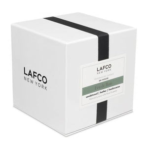 The LAFCO - FEU DE BOIS, SKI HOUSE 6.5OZ by LAFCO is a white, cylindrical candle encased in a clear glass holder with a slightly green-tinted top edge. Infused with soothing scents of frankincense and sandalwood, it casts a soft shadow to the right on a white background.