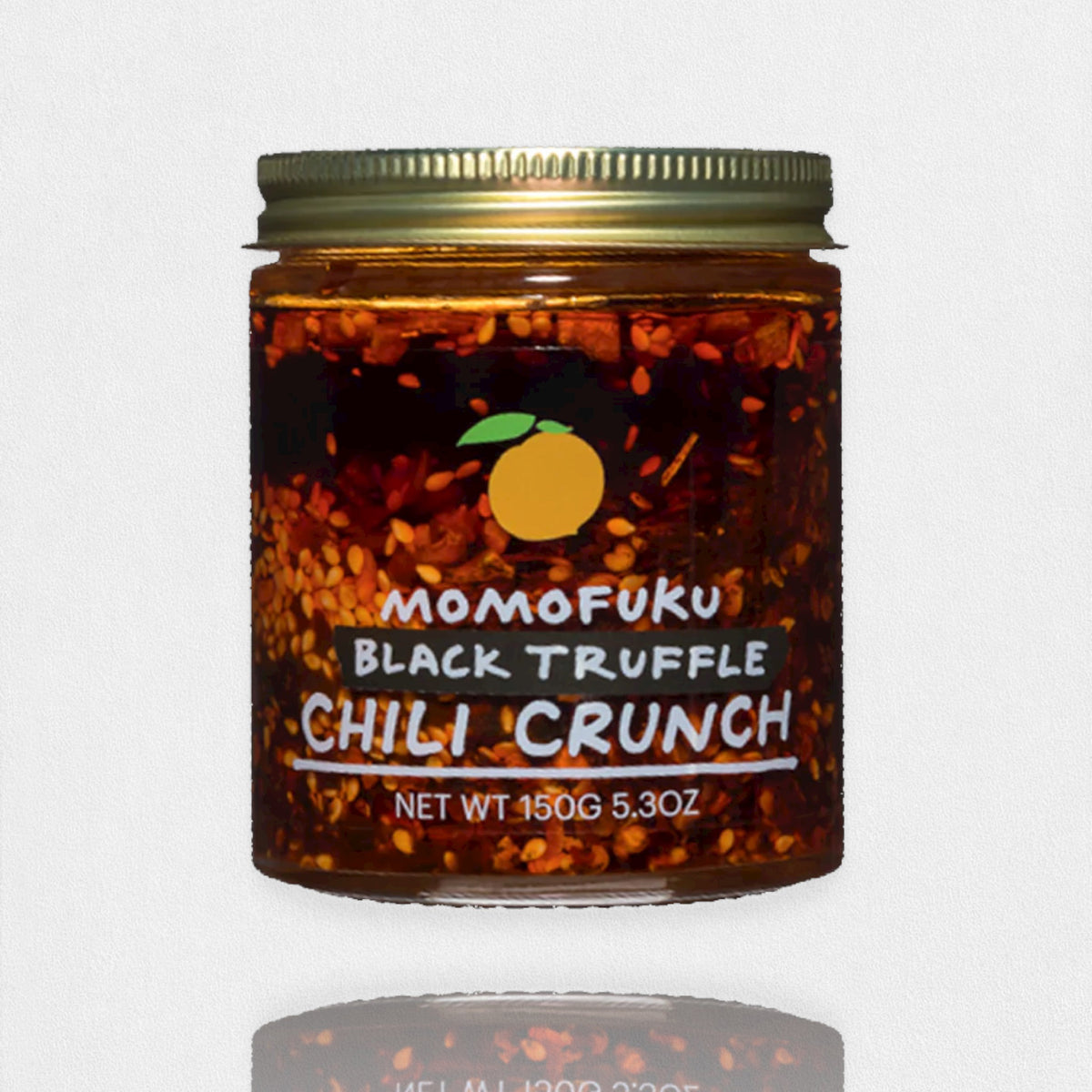 A jar of MOMOFUKU - BLACK TRUFFLE CHILI CRUNCH is shown against a plain background. The jar contains a mixture of chili flakes and oil, and features a label with an orange logo and text indicating "MOMOFUKU - BLACK TRUFFLE CHILI CRUNCH" alongside the net weight of 150g (5.3 oz).