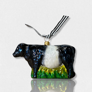 A luminous, hand-painted FEARRINGTON SIGNATURE COLLECTION - HANDMADE BELTIE ORNAMENT from JOY TO THE WORLD COLLECTIBLES dangles by a black and white striped ribbon. Shaped like a cow with a glossy surface that catches the light, it features a black head and rear with a white midsection. This delightful piece exudes the spirit of Fearrington Village, perched on a green base resembling grass.
