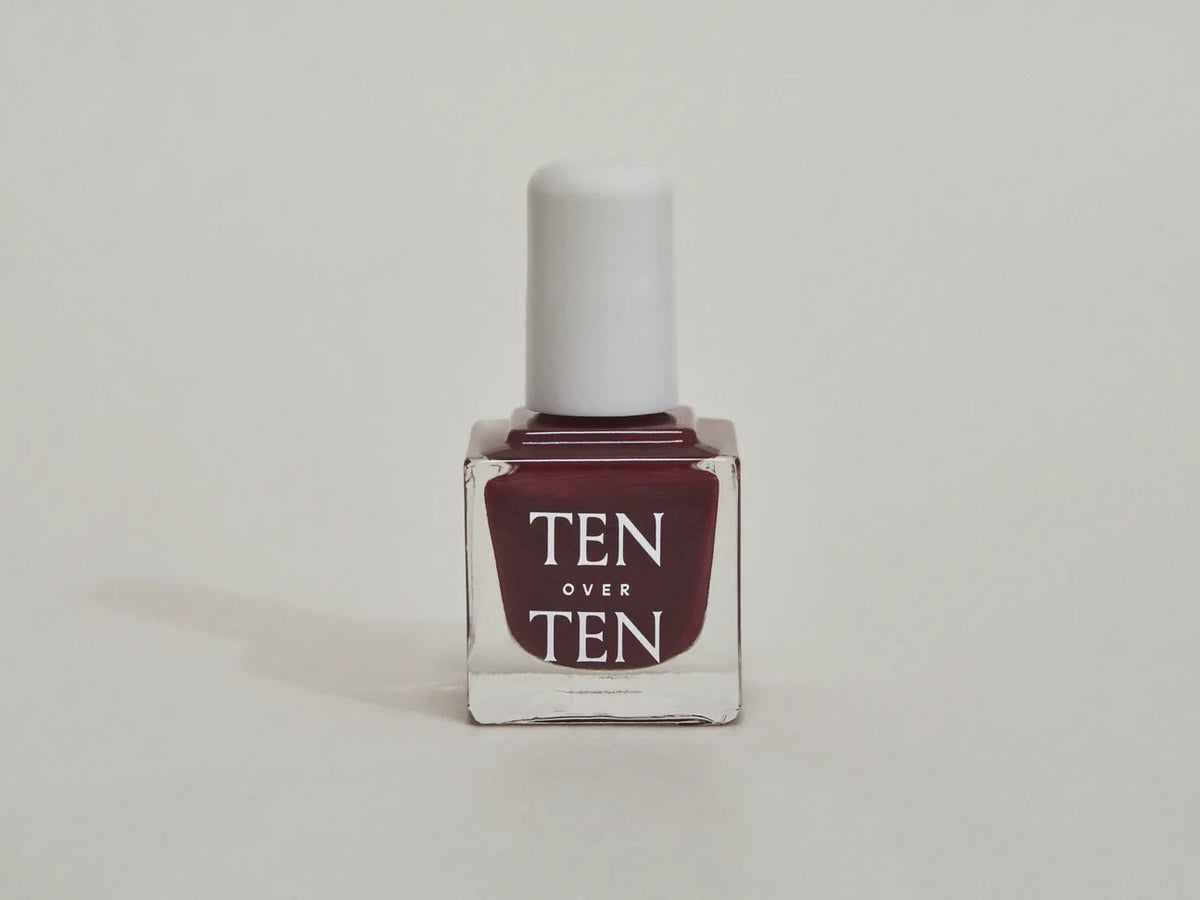 A glass bottle of TENOVERTEN - NAIL POLISH from the brand TEN OVER TEN, featuring a white cap and a dusty pink nail color. The front label displays "TEN OVER TEN" in white uppercase letters. This vegan, eight-free formula ensures both beauty and peace of mind. The background is plain and light-colored.