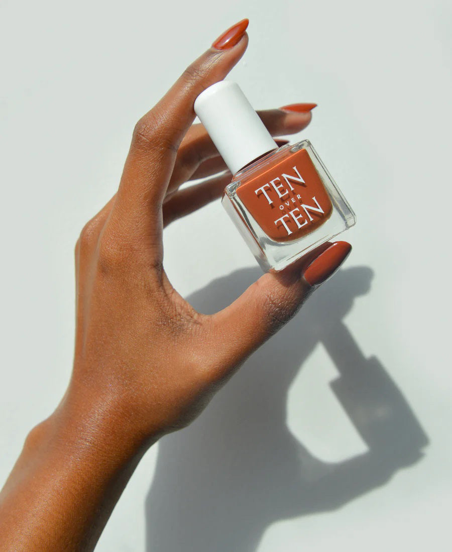A glass bottle of TENOVERTEN - NAIL POLISH from the brand TEN OVER TEN, featuring a white cap and a dusty pink nail color. The front label displays "TEN OVER TEN" in white uppercase letters. This vegan, eight-free formula ensures both beauty and peace of mind. The background is plain and light-colored.