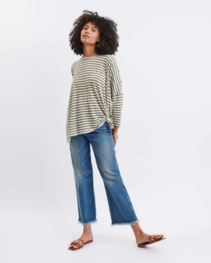 A woman with long, straight hair stands against a white wall, wearing the MERSEA Catalina Slub Tee in blue paired with white pants. The midweight textured cotton tee is from MERSEA. She looks at the camera with a neutral expression.