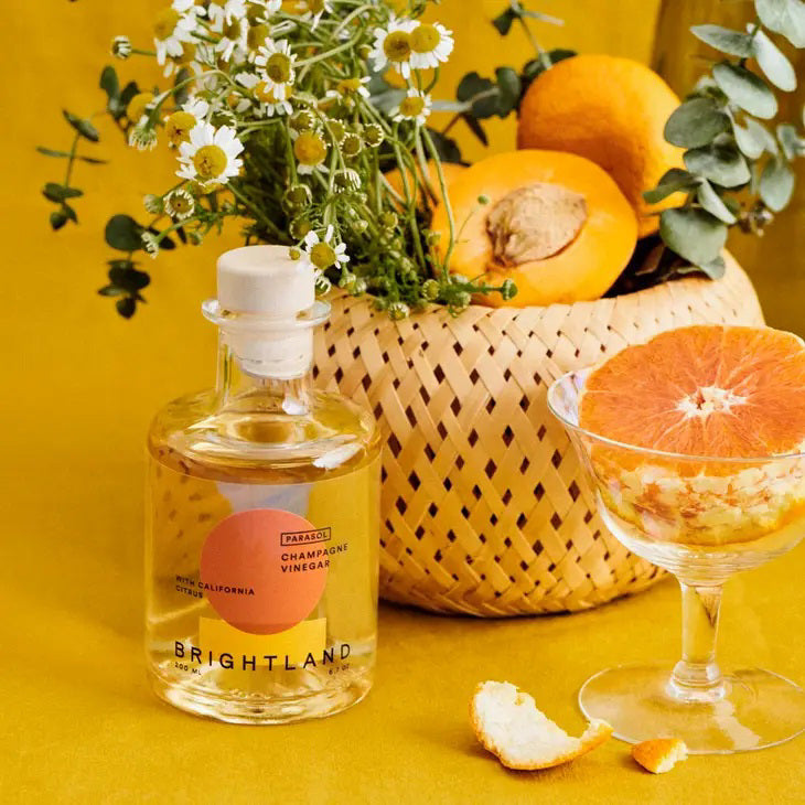 A clear glass bottle of Brightland &#39;Parasol&#39; Citrus Champagne Vinegar with a wooden stopper. The label features a bold design with an orange circle and yellow semicircle. Text on the label reads &quot;Parasol Champagne Vinegar with California Citrus, Navel and Valencia oranges, Brightland, 200 mL, 6.7 oz.