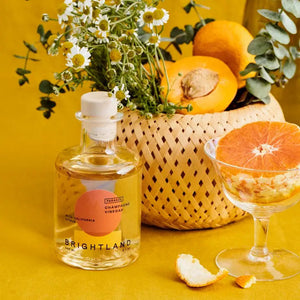 A clear glass bottle of Brightland 'Parasol' Citrus Champagne Vinegar with a wooden stopper. The label features a bold design with an orange circle and yellow semicircle. Text on the label reads "Parasol Champagne Vinegar with California Citrus, Navel and Valencia oranges, Brightland, 200 mL, 6.7 oz.
