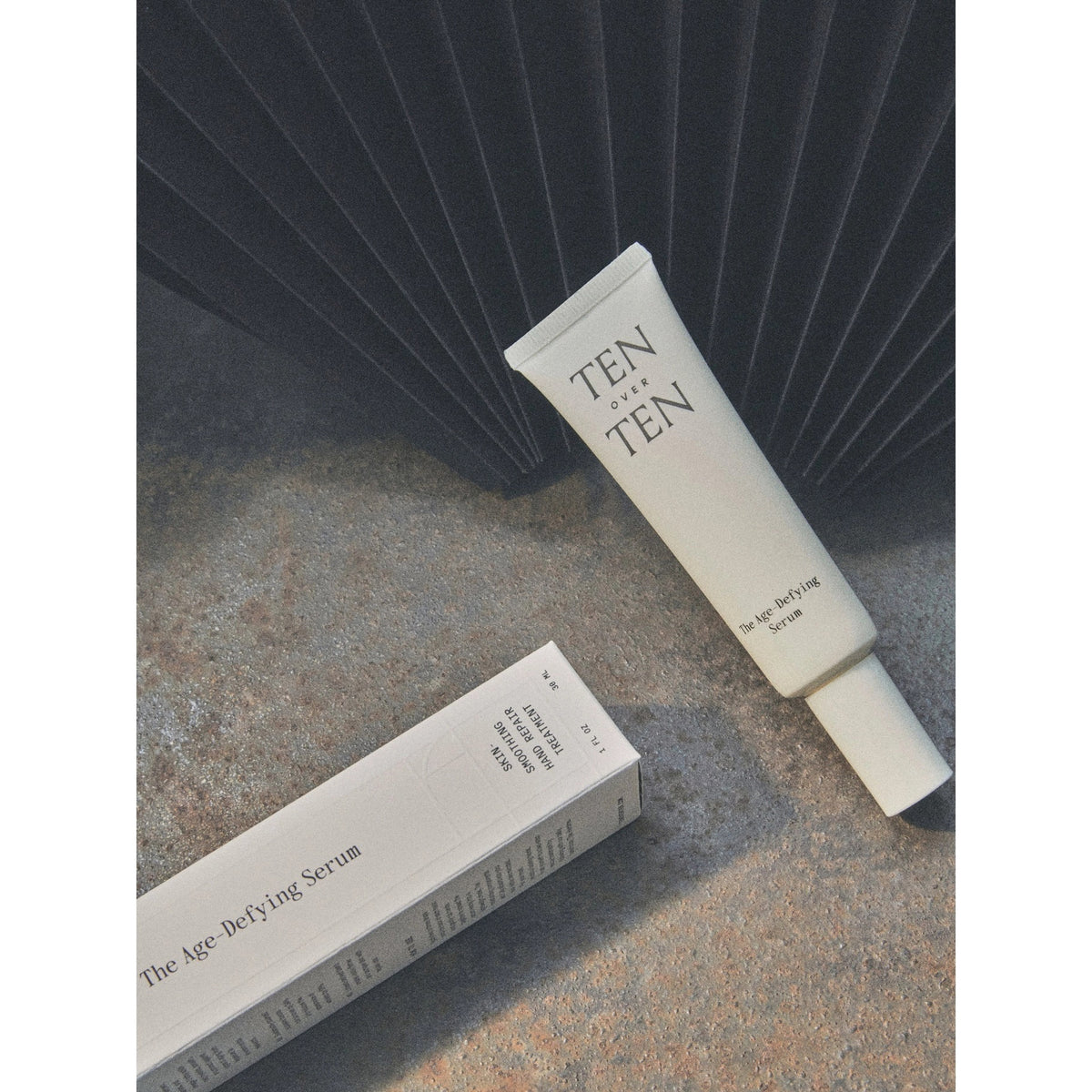 A tube labeled "TENOVERTEN - AGE DEFYING SERUM REPAIR TREATMENT" by TEN OVER TEN rests on a textured surface next to its box, with a dark fan-like object behind—ideal as an anti-aging and restorative hand serum.