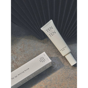 A tube labeled "TENOVERTEN - AGE DEFYING SERUM REPAIR TREATMENT" by TEN OVER TEN rests on a textured surface next to its box, with a dark fan-like object behind—ideal as an anti-aging and restorative hand serum.