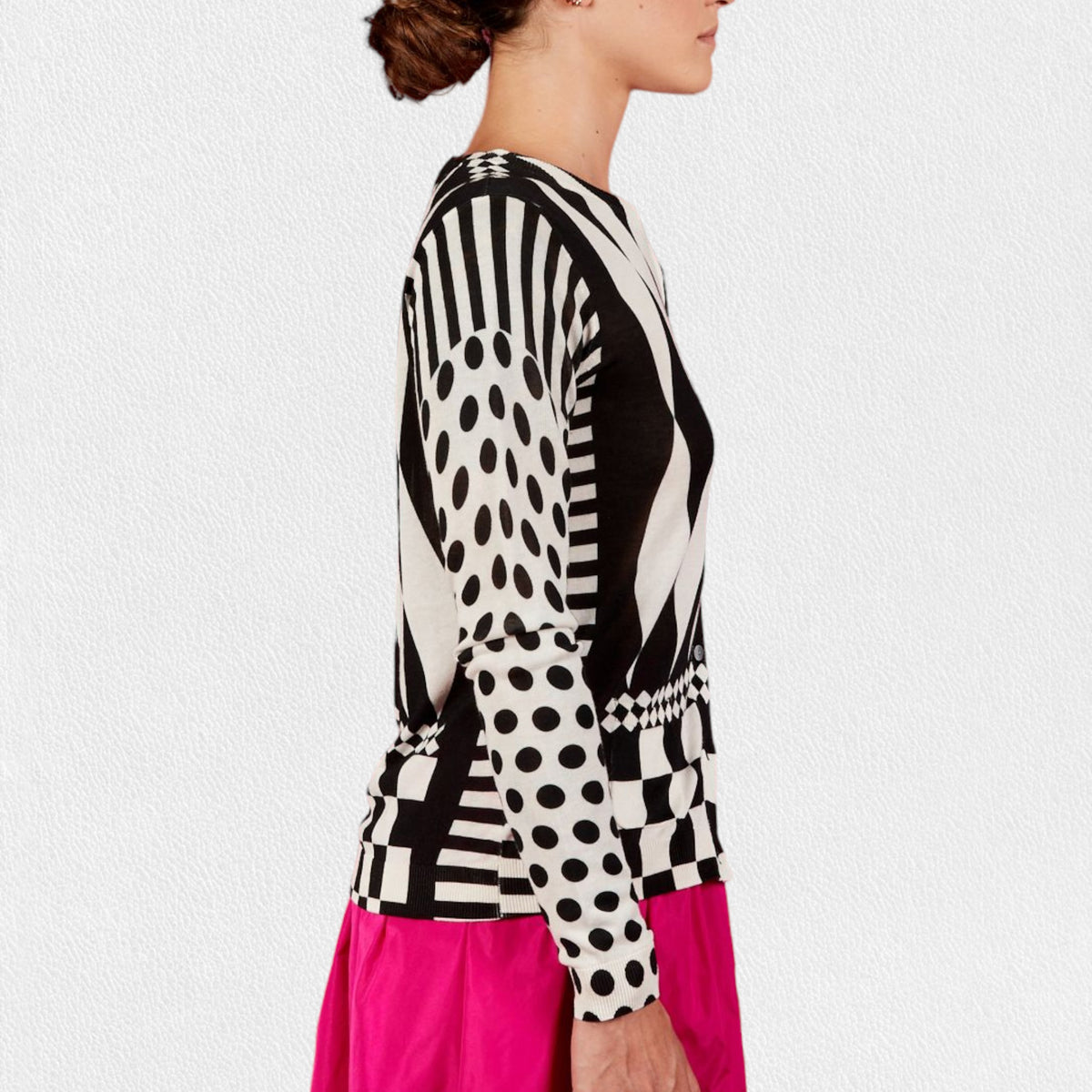 A person wearing the IN BED WITH YOU - GEOMETRIC PRINT CARDIGAN from the Italian brand IN BED WITH YOU/CLAUDIOLAI SRL, featuring alternating diamond shapes on the front, polka dots on the sleeves, and a checkerboard pattern at the hem. They are also wearing a bright pink skirt.
