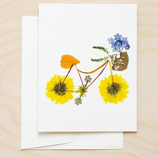 Introducing the PETAL PEOPLE PRESS - FLOWER PEDALS CARD from PETAL PEOPLE PRESS: a high-resolution digital print greeting card showcasing a bicycle masterfully crafted from pressed flowers and leaves. The design features large yellow flowers as wheels, a frame made from stems and leaves, and handlebar and basket adorned with clusters of small blue flowers and greenery. An envelope is included.