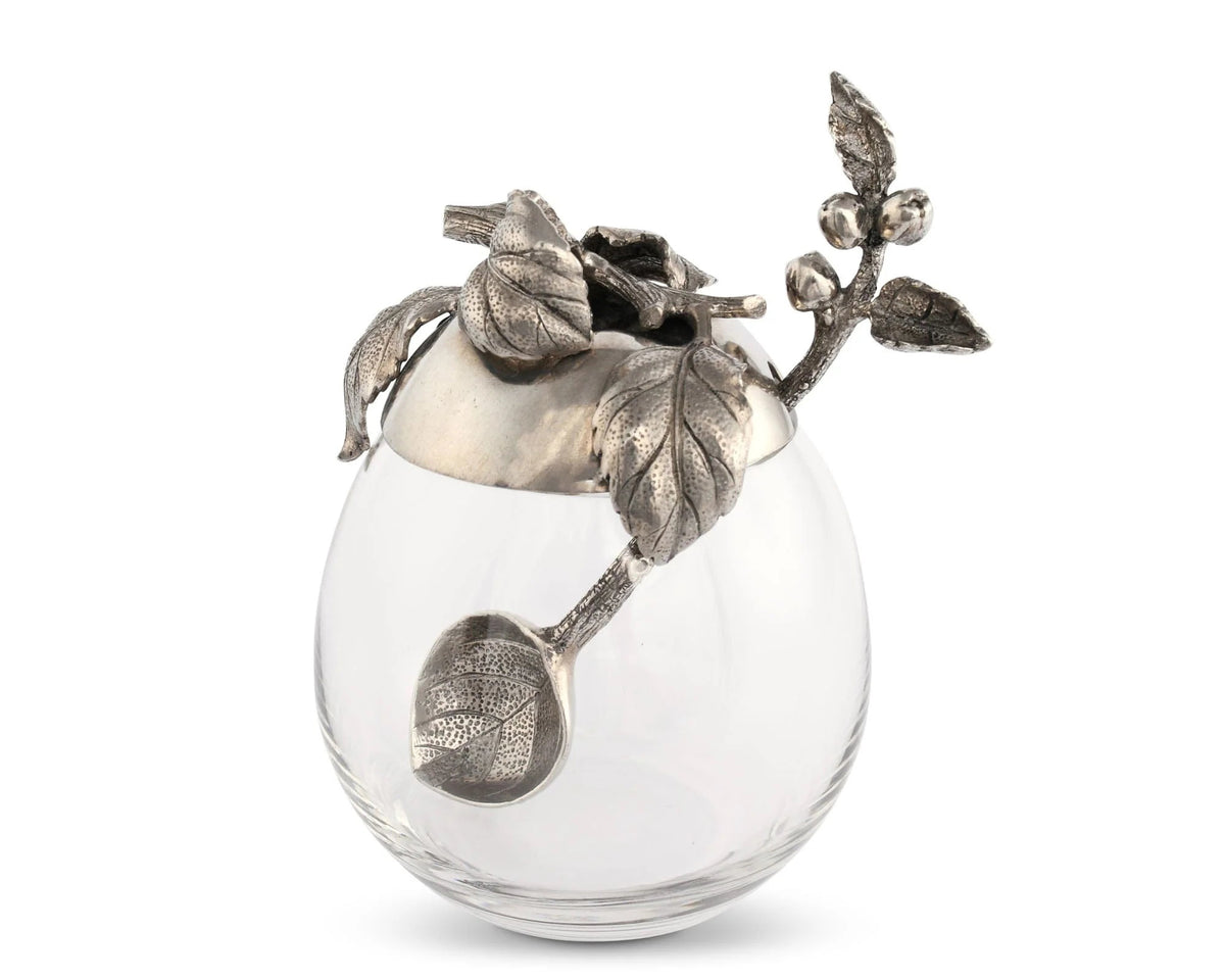 The VAGABOND HOUSE - APPLE HONEY POT features a decorative glass jar with a pewter cap showcasing intricate leaf and berry designs. The cap&#39;s handle mimics a stem with leaves, adding ornate elegance to this centerpiece by VAGABOND HOUSE, ideal for outdoor dining.