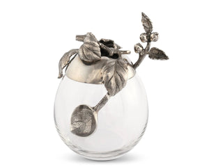 The VAGABOND HOUSE - APPLE HONEY POT features a decorative glass jar with a pewter cap showcasing intricate leaf and berry designs. The cap's handle mimics a stem with leaves, adding ornate elegance to this centerpiece by VAGABOND HOUSE, ideal for outdoor dining.