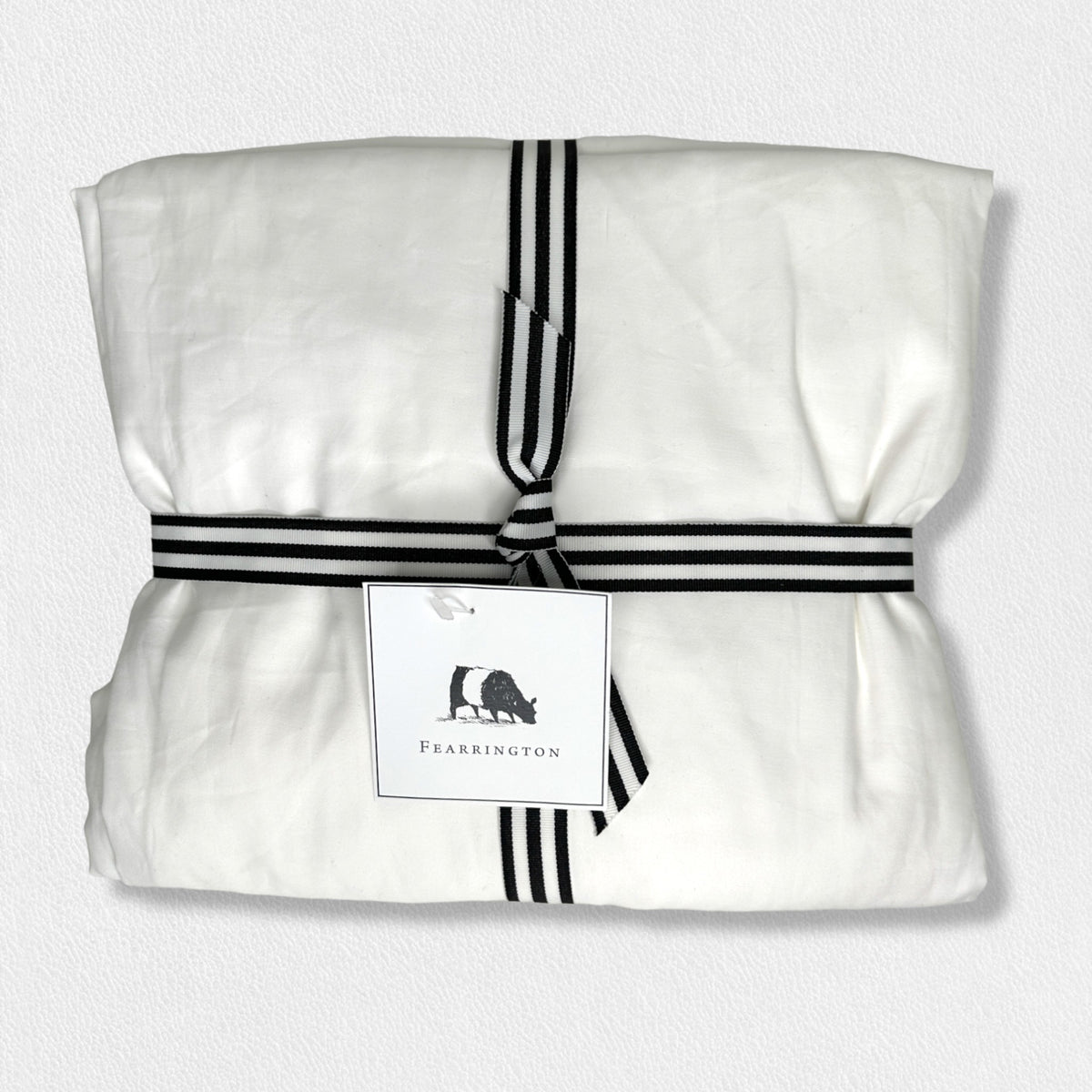 A neatly folded white fabric bundle of luxury bedding tied with a black and white striped ribbon, featuring a white tag with a black silhouette of a farm animal and the text &quot;Fearrington.&quot; This Frette North America Fearrington Lifestyle Bedding Collection&#39;s Frette King Fitted Sheets boasts a 300 thread count, set against a light gray textured background.