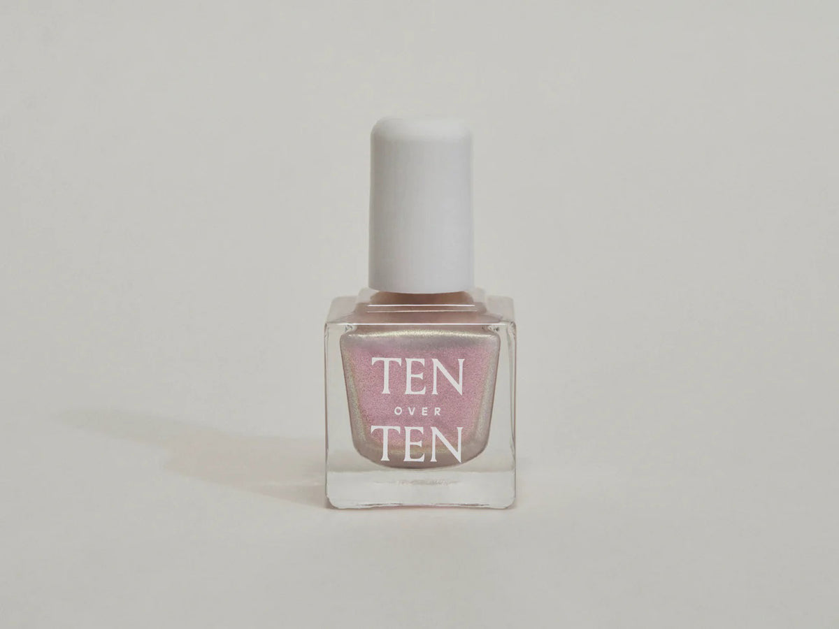 A glass bottle of TENOVERTEN - NAIL POLISH from the brand TEN OVER TEN, featuring a white cap and a dusty pink nail color. The front label displays "TEN OVER TEN" in white uppercase letters. This vegan, eight-free formula ensures both beauty and peace of mind. The background is plain and light-colored.