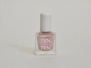 A glass bottle of TENOVERTEN - NAIL POLISH from the brand TEN OVER TEN, featuring a white cap and a dusty pink nail color. The front label displays "TEN OVER TEN" in white uppercase letters. This vegan, eight-free formula ensures both beauty and peace of mind. The background is plain and light-colored.