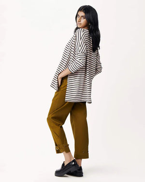 A woman with long, straight hair stands against a white wall, wearing the MERSEA Catalina Slub Tee in blue paired with white pants. The midweight textured cotton tee is from MERSEA. She looks at the camera with a neutral expression.