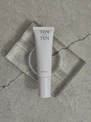 A beige tube labeled "TENOVERTEN - THE HEROINE HAND LOTION" by TEN OVER TEN rests on a clear rectangular platform atop a textured, cracked concrete surface.