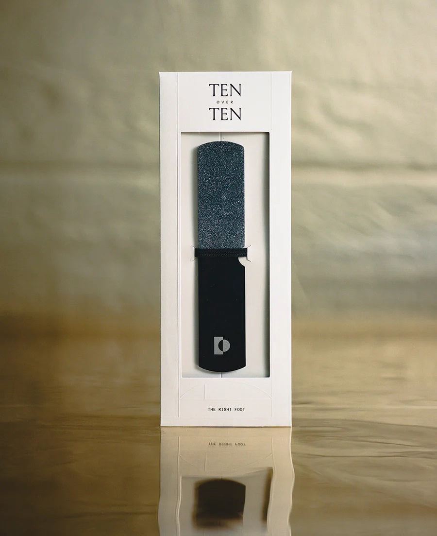 The TENOVERTEN - THE RIGHT FOOT NAIL FILE by TEN OVER TEN is a salon-grade black stainless steel file with a diamond-ceramic layer, showcased upright in white packaging with a window revealing the file against a gold surface.