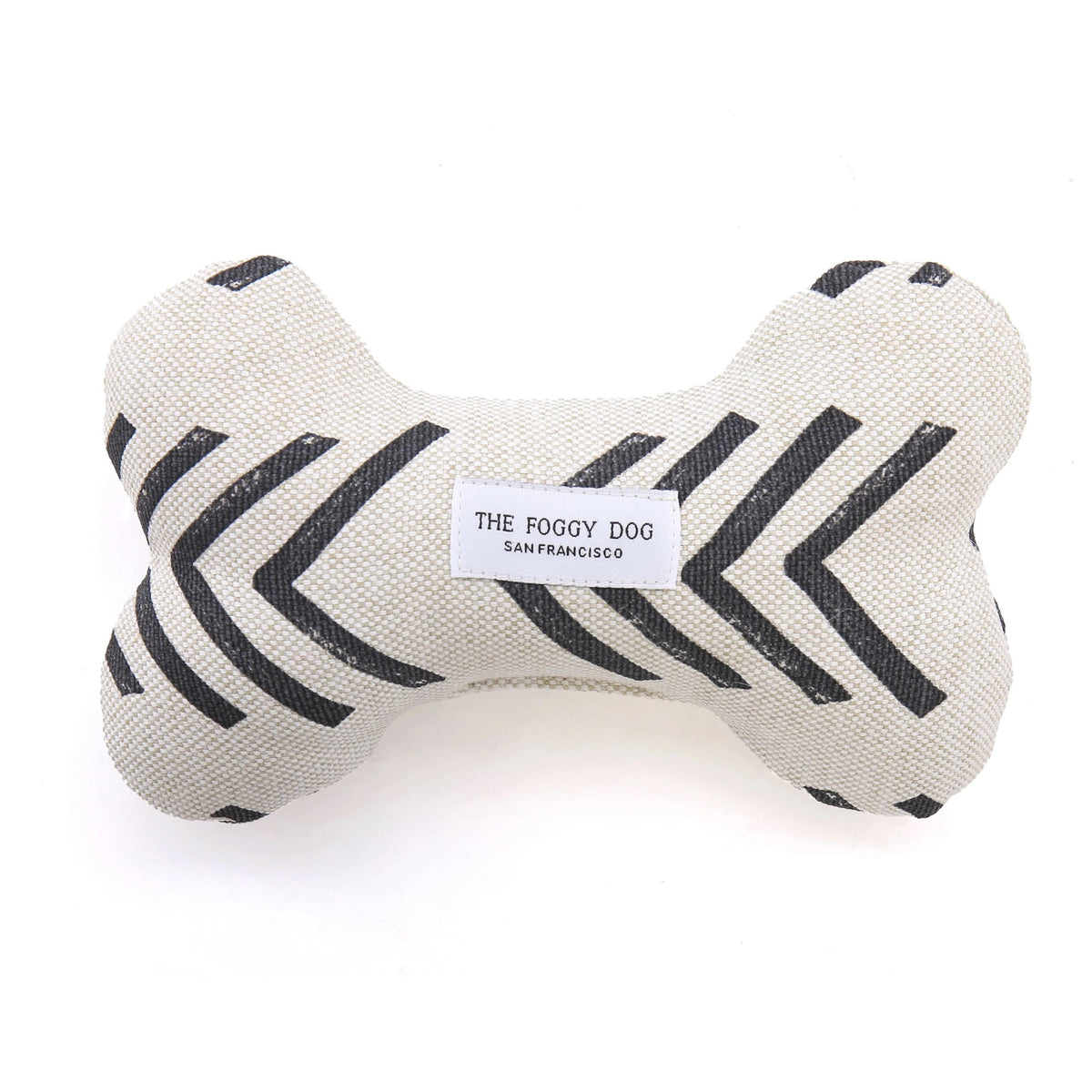 The DOG BONE SQUEAKY TOY (MODERN MUD CLOTH) by THE FOGGY DOG is a handcrafted canvas dog toy, featuring a bone shape made of beige fabric adorned with black zigzag patterns. This eco-friendly squeaker toy has a label in the center reading &quot;The Foggy Dog SAN FRANCISCO,&quot; filled with safe, eco-friendly fiberfill for enjoyable playtime, and is proudly made in the USA.