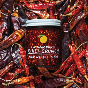 A jar of MOMOFUKU - CHILI CRUNCH, featuring a white lid filled with chili oil and visible chili flakes. The label showcases an orange fruit graphic and text that reads "MOMOFUKU - CHILI CRUNCH, Net Wt 150g (5.3oz)." Made with umami-rich ingredients, it's perfect for fans of Chinese chili crisp sauces. The background is plain white.