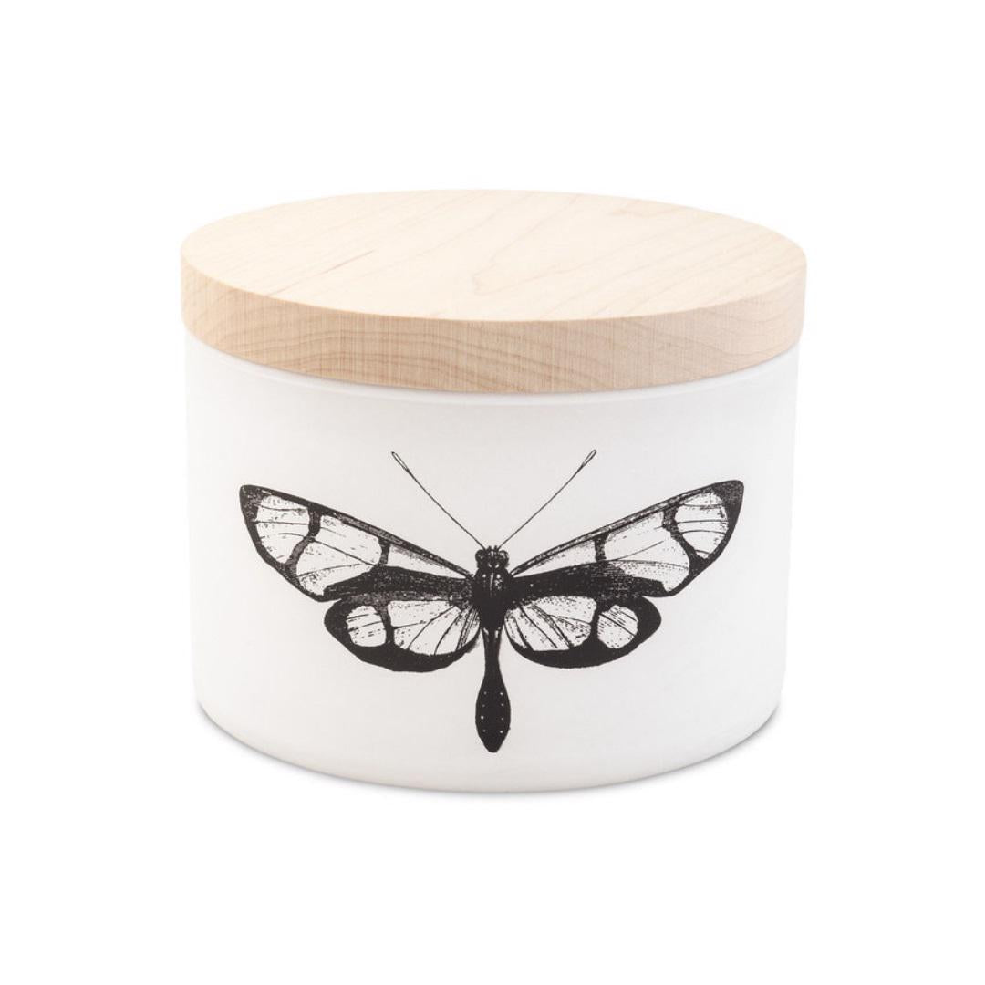 The CITRONELLA EUCALYPTUS 3 WICK CANDLE by SKEEM CANDLES is presented in a white ceramic jar topped with a light wooden lid. Adorning the front is an intricate black and white butterfly illustration. Inside, this sophisticated container holds a triple wick candle infused with Citronella oil, ideal for repelling insects.