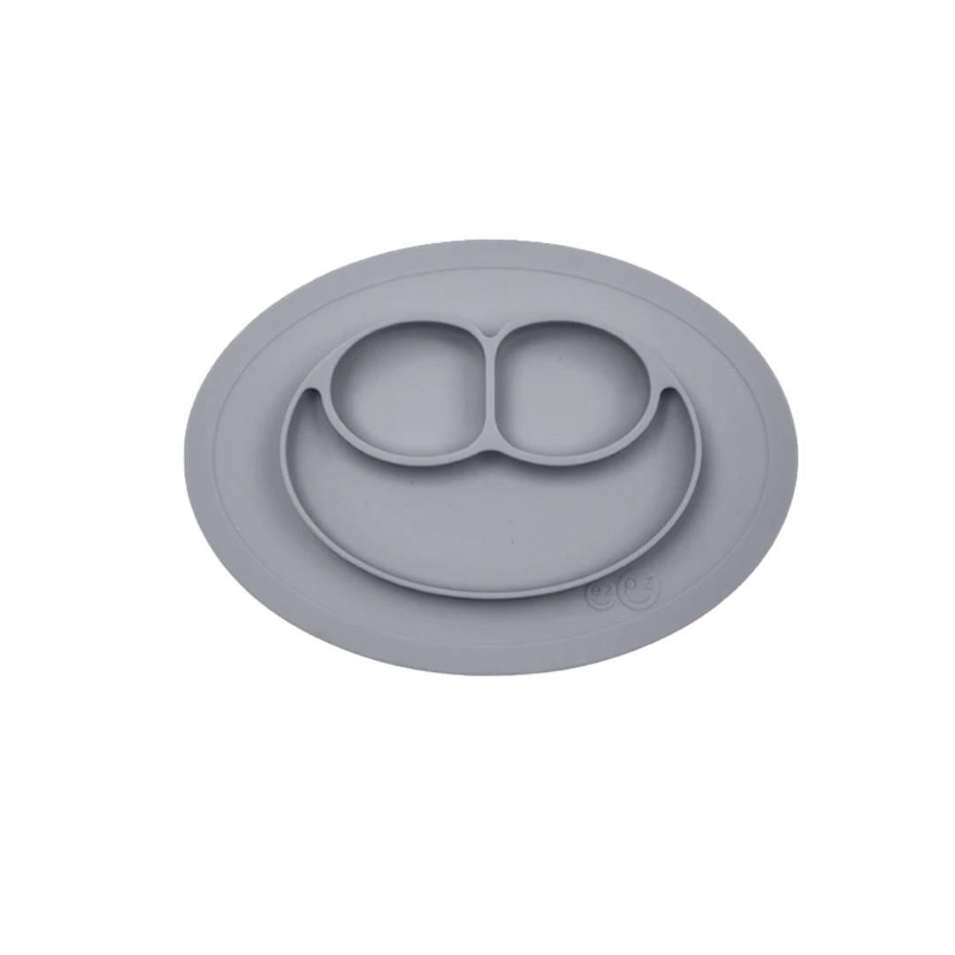 The EZPZ MINI MAT (PEWTER) is a gray silicone baby plate shaped like a smiling face, featuring three compartments for easy self-feeding. Ideal as a feeding mat, it's shown against a white background.
