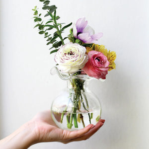 The VIETRI - HIBISCUS BUD VASE CLEAR by VIETRI features an artistic, wavy, and ruffled opening with a round base. This design resembles a blooming flower, crafted from smooth, transparent glass to showcase a modern and elegant aesthetic, making it perfect for any floral arrangement.