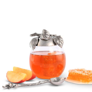 The VAGABOND HOUSE - APPLE HONEY POT features a decorative glass jar with a pewter cap showcasing intricate leaf and berry designs. The cap's handle mimics a stem with leaves, adding ornate elegance to this centerpiece by VAGABOND HOUSE, ideal for outdoor dining.