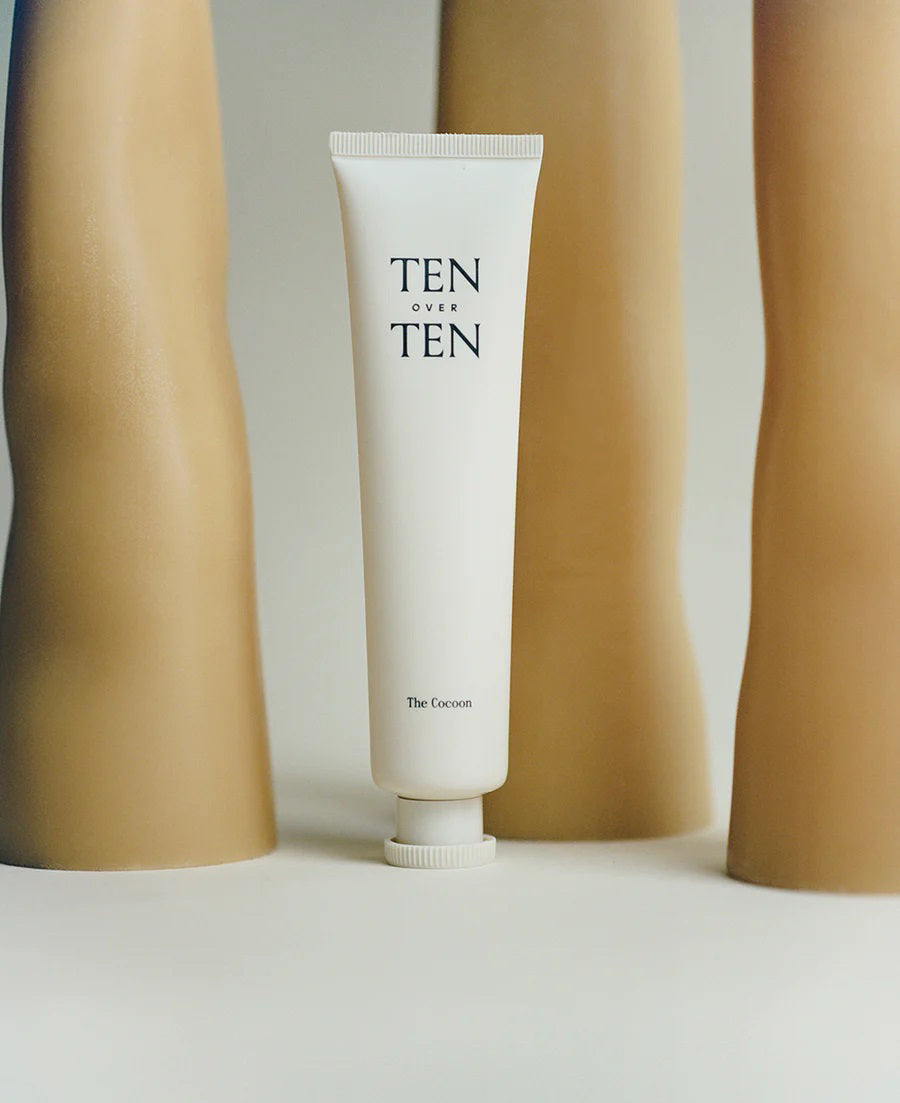 Two people, one with bare shoulders and another in white, hold a TENOVERTEN - THE COCOON HAND &amp; FOOT CREAM tube by TEN OVER TEN—suggesting a plant-based cream for long-lasting moisture on dry hands and feet.