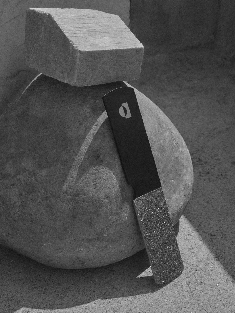 The TENOVERTEN - THE RIGHT FOOT NAIL FILE by TEN OVER TEN is a salon-grade black stainless steel file with a diamond-ceramic layer, showcased upright in white packaging with a window revealing the file against a gold surface.