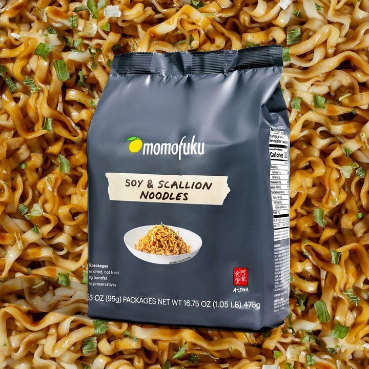 MOMOFUKU Soy and Scallion Noodles come in dark gray packaging with white and yellow accents, featuring a bowl of air-dried noodles on the front. Each pack highlights five servings, no preservatives, and restaurant-level flavor.