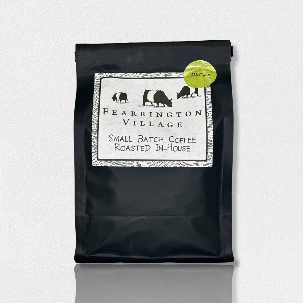 A black package of Belted Goat Fearrington Signature Collection Small Batch House Blended Decaf Coffee Beans. The label features three black animal silhouettes and a decorative border. A green sticker in the upper-right corner indicates the Fearrington House decaf blend, made from Brazilian and Colombian beans. The background is textured white.