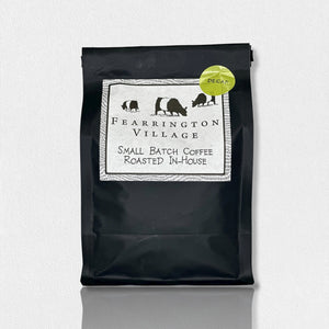 A black package of Belted Goat Fearrington Signature Collection Small Batch House Blended Decaf Coffee Beans. The label features three black animal silhouettes and a decorative border. A green sticker in the upper-right corner indicates the Fearrington House decaf blend, made from Brazilian and Colombian beans. The background is textured white.