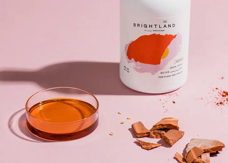 A white bottle with a cork lid, labeled "BRIGHTLAND" in black text. The label also features an orange-red abstract shape with the words "100% Chili Olive Oil" and notes "For flame and fervor." Made from Heirloom Frantoio Olives, this minimalist design stands against a plain background.