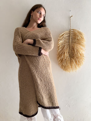 A person with long hair stands against a beige wall, wearing the oversized, cozy ROSE LONG WOOL DUSTER by DEBBIE KATZ, featuring brown trim at the sleeves and hem. They have one arm folded across their waist. A decorative, large dried grass wall hanging is visible on the right.