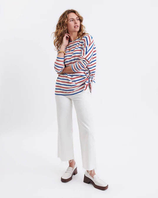 A woman with long, straight hair stands against a white wall, wearing the MERSEA Catalina Slub Tee in blue paired with white pants. The midweight textured cotton tee is from MERSEA. She looks at the camera with a neutral expression.