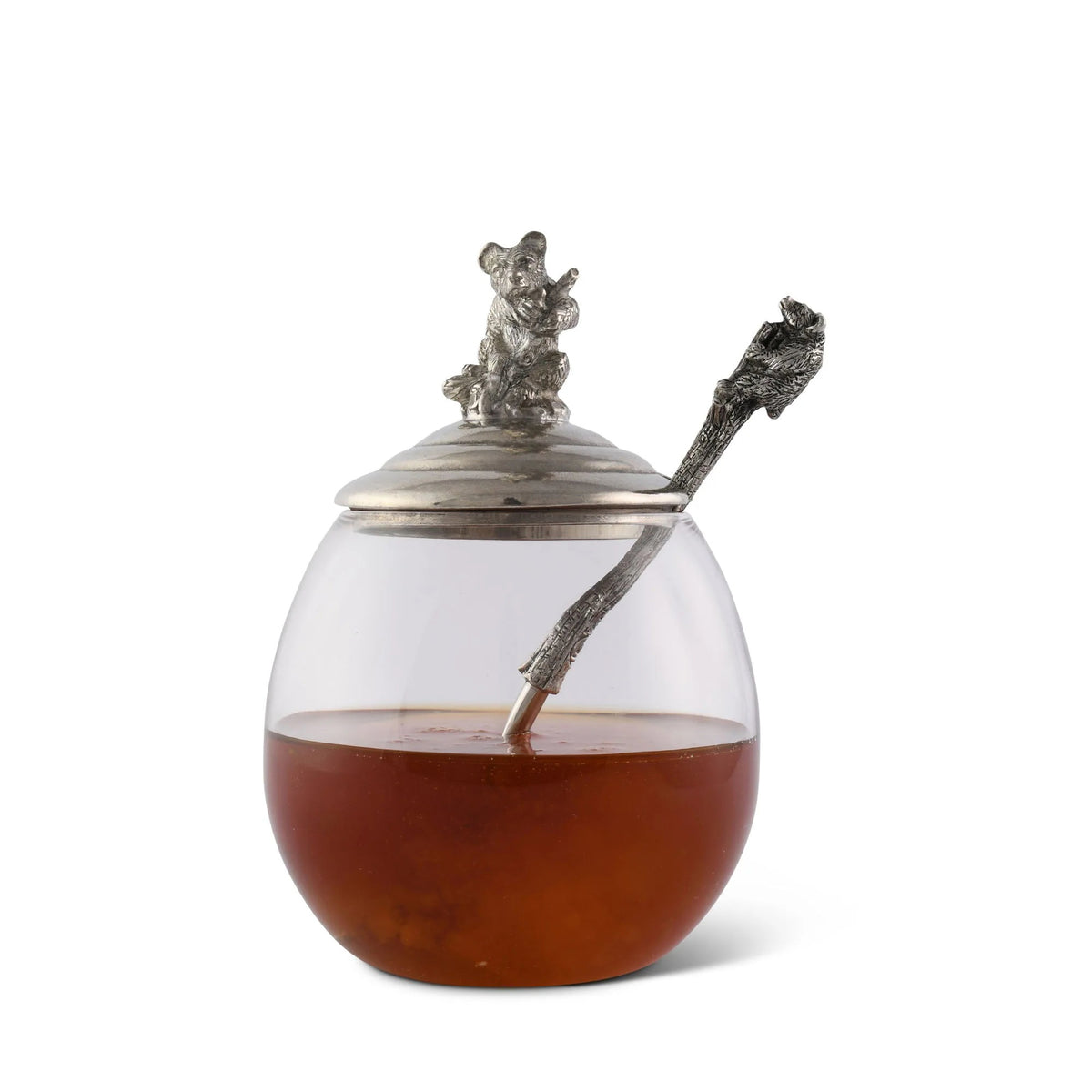 The VAGABOND HOUSE - LITTLE BEAR HONEY POT is a clear glass jar with a luxury pewter lid featuring a bear on top and includes a tree branch-styled silver spoon. This elegant yet rustic design by VAGABOND HOUSE resembles a little bear honey pot.