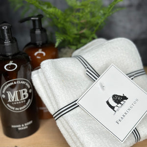 A neatly folded white textured towel, part of the FEARRINGTON LIFESTYLE TOWEL COLLECTION - WAFFLE FACE CLOTH S/4 by HARBOR LINEN, is tied with a black and white striped ribbon. Attached to the ribbon is a square tag featuring a black silhouette of a cow with "FEARRINGTON" in capital letters beneath it. The towel sits on a white surface.