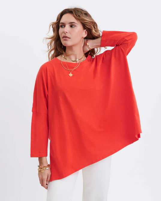 A woman with long, straight hair stands against a white wall, wearing the MERSEA Catalina Slub Tee in blue paired with white pants. The midweight textured cotton tee is from MERSEA. She looks at the camera with a neutral expression.