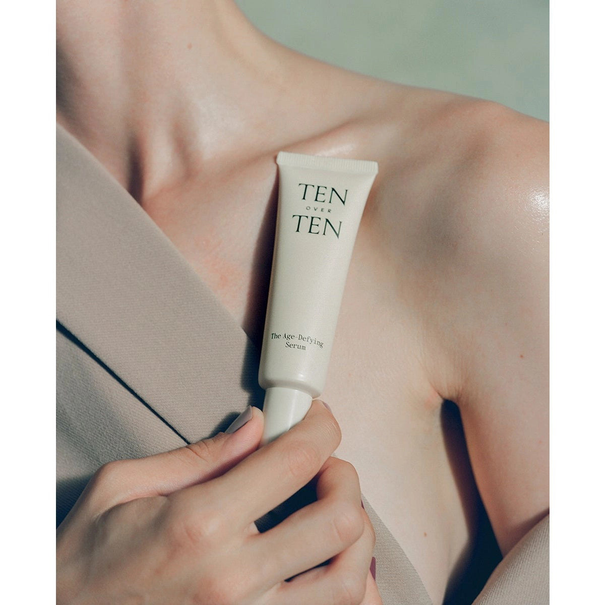 A tube labeled "TENOVERTEN - AGE DEFYING SERUM REPAIR TREATMENT" by TEN OVER TEN rests on a textured surface next to its box, with a dark fan-like object behind—ideal as an anti-aging and restorative hand serum.