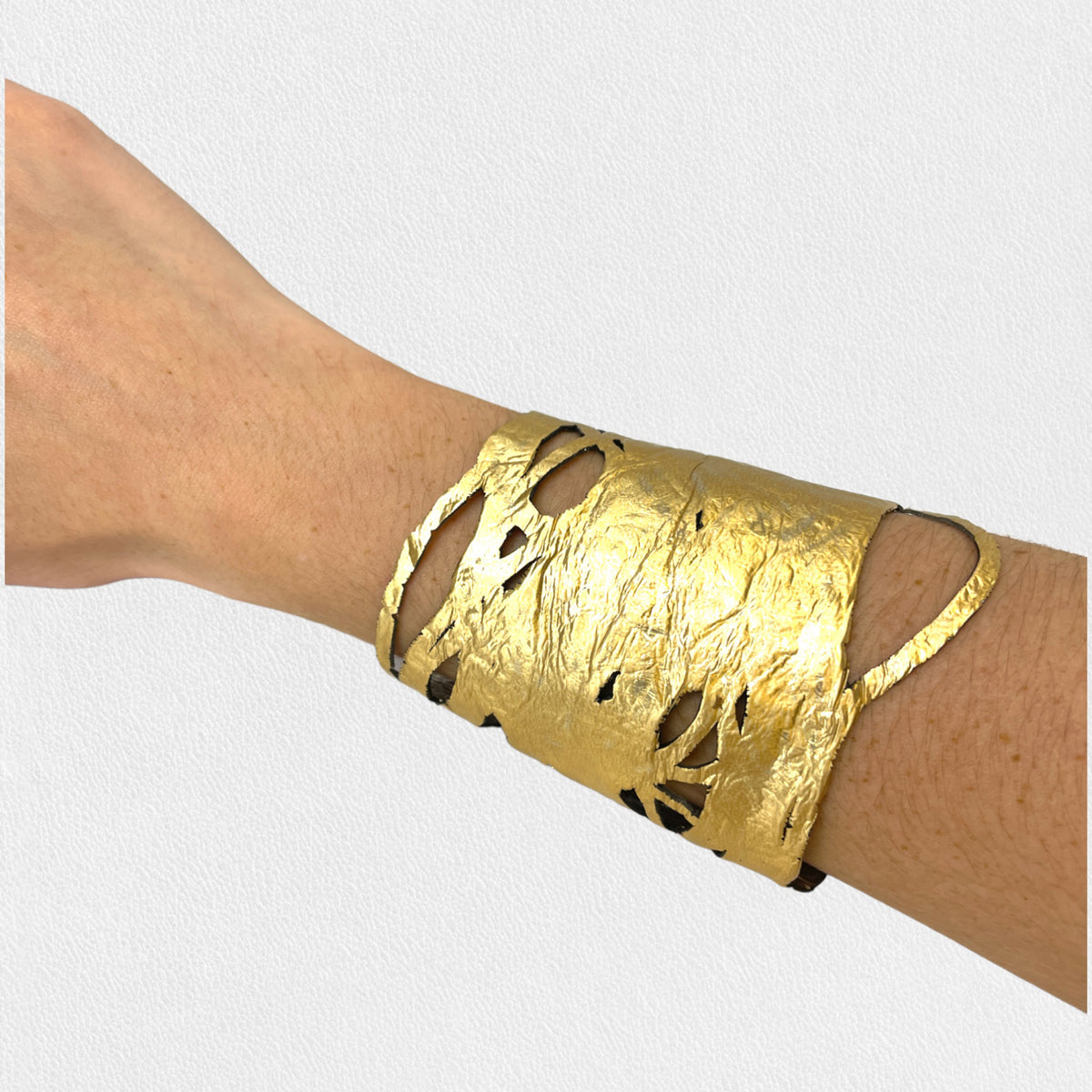 A person wearing the ILLUSIONISM BRACELET IN GOLD AND SILVER (REVERSIBLE) from SI SABATO ISABEL on their wrist. The handmade jewellery, crafted by an Argentinian artist, features irregular cut-out shapes and an intricate, hammered surface. The person is dressed in a faded blue denim shirt with white buttons.