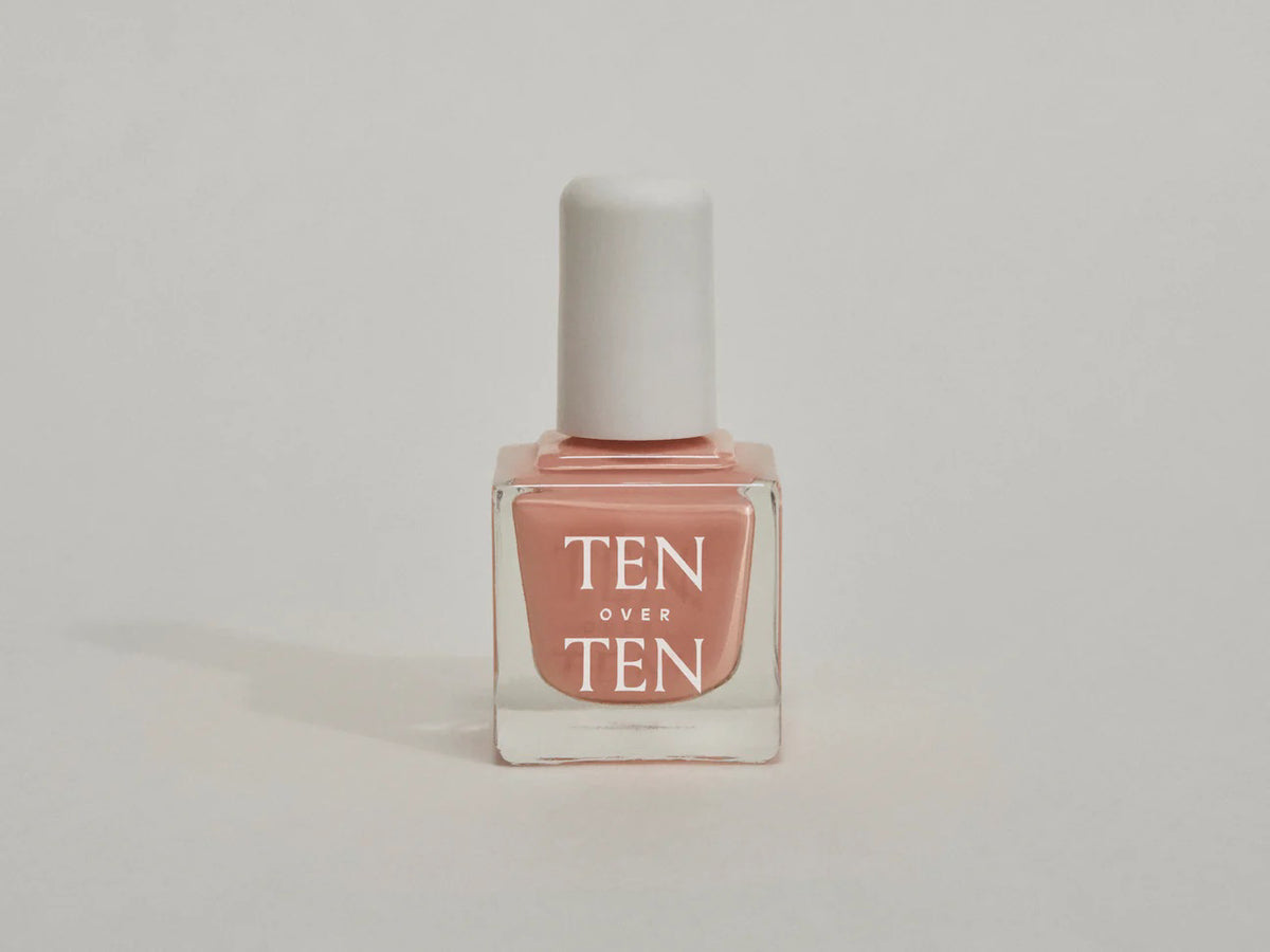 A glass bottle of TENOVERTEN - NAIL POLISH from the brand TEN OVER TEN, featuring a white cap and a dusty pink nail color. The front label displays "TEN OVER TEN" in white uppercase letters. This vegan, eight-free formula ensures both beauty and peace of mind. The background is plain and light-colored.