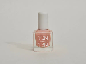 A glass bottle of TENOVERTEN - NAIL POLISH from the brand TEN OVER TEN, featuring a white cap and a dusty pink nail color. The front label displays "TEN OVER TEN" in white uppercase letters. This vegan, eight-free formula ensures both beauty and peace of mind. The background is plain and light-colored.