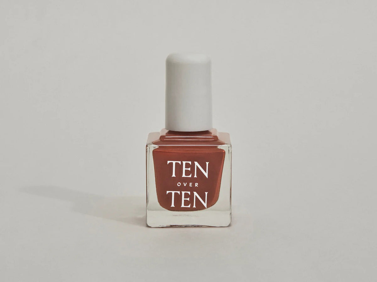 A glass bottle of TENOVERTEN - NAIL POLISH from the brand TEN OVER TEN, featuring a white cap and a dusty pink nail color. The front label displays "TEN OVER TEN" in white uppercase letters. This vegan, eight-free formula ensures both beauty and peace of mind. The background is plain and light-colored.