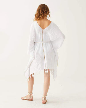 A woman with wavy hair smiles in the MERSEA - BEACH KAFTAN, a loose white Turkish cotton cover-up with fringe and gold trim, tied at the waist. She pairs it with sandals, showcasing MERSEA’s effortless resort style against a white background.