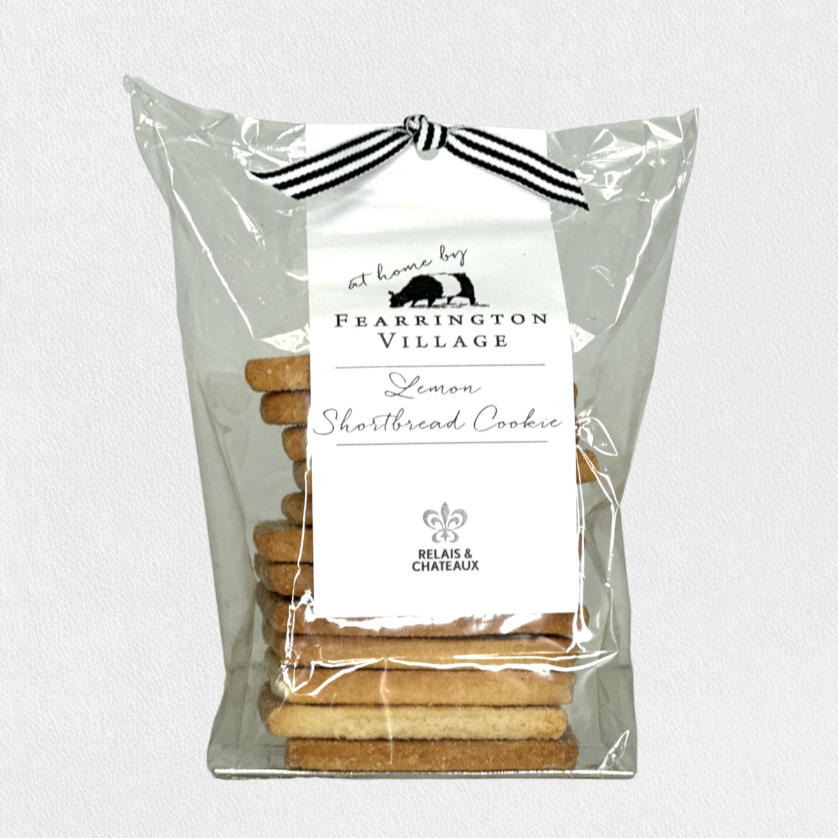 A stack of buttery, crisp FEARRINGTON SIGNATURE COLLECTION - HOUSEMADE LEMON SHORTBREAD COOKIES stands on a black round surface. Behind the stack, a clear plastic bag containing more cookies is tied with a black and white ribbon and features a label reading &quot;FEARRINGTON HOUSE Lemon Shortbread Cookie&quot; with decorative details from The Fearrington House Restaurant.
