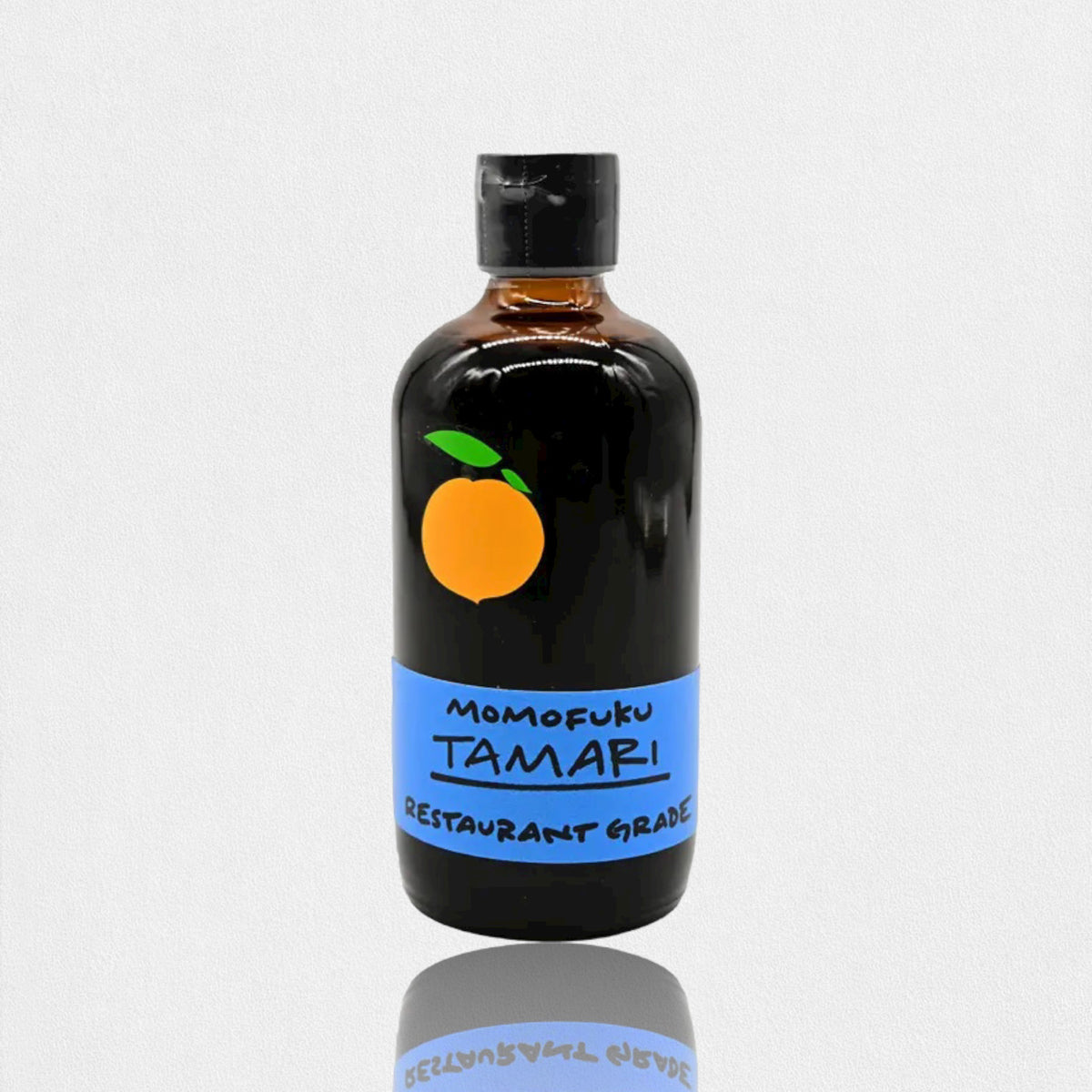 A brown glass bottle with a black cap contains MOMOFUKU - TAMARI, a restaurant-grade, gluten-free soy sauce made from organic ingredients. The label is blue with a minimalist orange fruit logo. The bottle is reflected on the white surface it sits on.