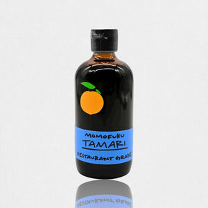 A brown glass bottle with a black cap contains MOMOFUKU - TAMARI, a restaurant-grade, gluten-free soy sauce made from organic ingredients. The label is blue with a minimalist orange fruit logo. The bottle is reflected on the white surface it sits on.